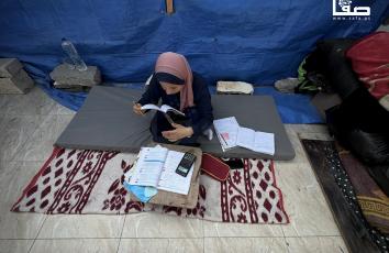 سارة قنن (17 عامًا) تراجع دروسها عن بُعد داخل خيمتها في خانيونس استعدادًا لامتحانات الثانوية العامة